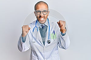 Hispanic adult man wearing doctor uniform and stethoscope angry and mad raising fists frustrated and furious while shouting with