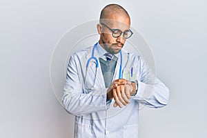 Hispanic adult man wearing doctor uniform and stethoscope checking the time on wrist watch, relaxed and confident