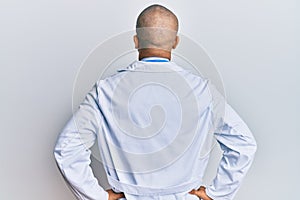 Hispanic adult man wearing doctor uniform and stethoscope standing backwards looking away with arms on body