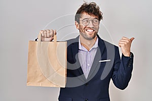 Hispanic business man holding delivery bag pointing thumb up to the side smiling happy with open mouth