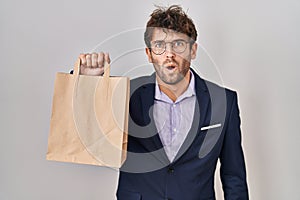 Hispanic business man holding delivery bag scared and amazed with open mouth for surprise, disbelief face