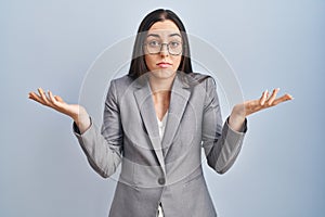 Hispanic business woman wearing glasses clueless and confused expression with arms and hands raised