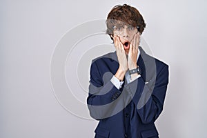 Hispanic business young man wearing glasses afraid and shocked, surprise and amazed expression with hands on face