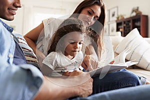 Hispanic couple and their young daughter sitting on the sofa reading a book together at home