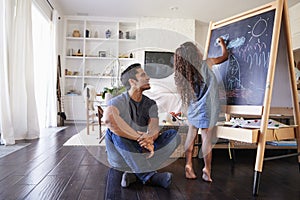 Hispanic dad sitting on the floor in sitting room watching his young daughter drawing on blackboard