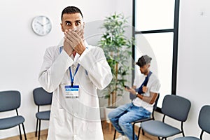 Hispanic doctor man at waiting room with pacient with arm injury shocked covering mouth with hands for mistake