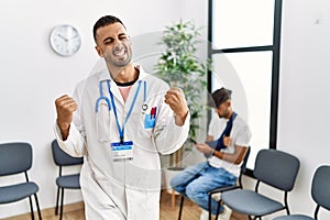 Hispanic doctor man at waiting room with pacient with arm injury very happy and excited doing winner gesture with arms raised,