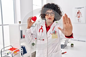 Hispanic doctor woman with curly hair holding heart with open hand doing stop sign with serious and confident expression, defense