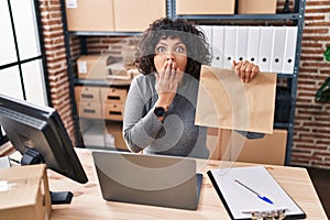 Hispanic doctor woman with curly hair working at small business ecommerce holding paper bag covering mouth with hand, shocked and