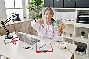 Hispanic doctor woman holding brazilian reals banknotes with angry face, negative sign showing dislike with thumbs down, rejection