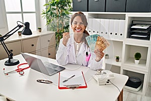 Hispanic doctor woman holding brazilian reals banknotes screaming proud, celebrating victory and success very excited with raised