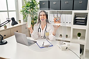 Hispanic doctor woman holding model of human anatomical skin and hair angry and mad screaming frustrated and furious, shouting photo