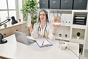 Hispanic doctor woman holding model of human anatomical skin and hair angry and mad screaming frustrated and furious, shouting photo