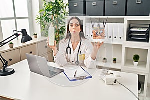 Hispanic doctor woman holding model of human anatomical skin and hair making fish face with mouth and squinting eyes, crazy and photo