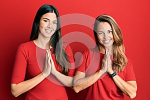 Hispanic family of mother and daughter wearing casual clothes over red background praying with hands together asking for