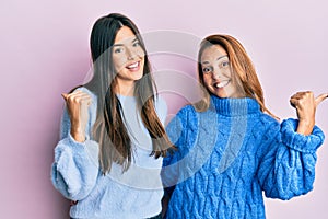 Hispanic family of mother and daughter wearing wool winter sweater pointing to the back behind with hand and thumbs up, smiling