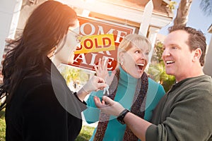 Hispanic Female Real Estate Agent Handing Over New House Keys to Happy Couple In Front of Sold For Sale Real Estate Sign