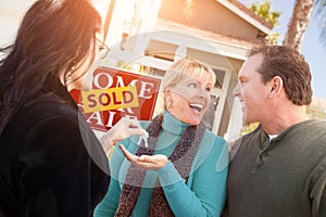 Hispanic Female Real Estate Agent Handing Over New House Keys to Happy Couple In Front of Sold For Sale Real Estate Sign