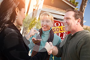 Hispanic Female Real Estate Agent Handing Over New House Keys to Happy Couple In Front of Sold For Sale Real Estate Sign