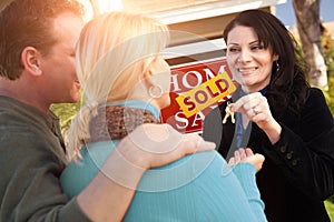 Hispanic Female Real Estate Agent Handing Over New House Keys to Happy Couple In Front of Sold For Sale Real Estate Sign