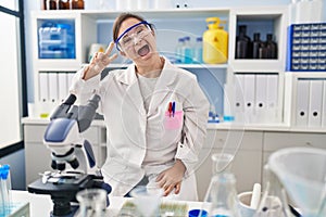 Hispanic girl with down syndrome working at scientist laboratory doing peace symbol with fingers over face, smiling cheerful
