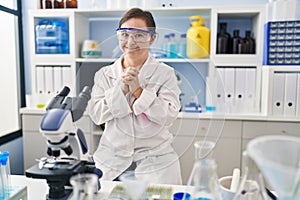 Hispanic girl with down syndrome working at scientist laboratory hands together and fingers crossed smiling relaxed and cheerful