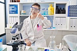 Hispanic girl with down syndrome working at scientist laboratory pointing with hand finger to face and nose, smiling cheerful