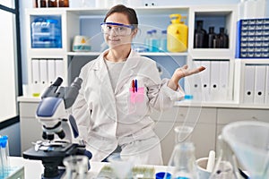 Hispanic girl with down syndrome working at scientist laboratory smiling cheerful presenting and pointing with palm of hand