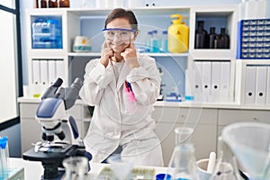 Hispanic girl with down syndrome working at scientist laboratory smiling with open mouth, fingers pointing and forcing cheerful