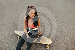 Hispanic girl with longboard sitting on the street and looking down. Portrait of stylish brunette girl with curly hair sits on a