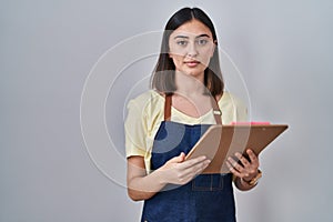 Hispanic girl wearing apron holding clipboard thinking attitude and sober expression looking self confident