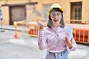 Hispanic girl wearing architect hardhat at construction site pointing thumb up to the side smiling happy with open mouth