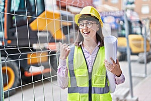 Hispanic girl wearing architect hardhat at construction site pointing thumb up to the side smiling happy with open mouth