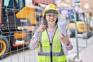 Hispanic girl wearing architect hardhat at construction site smiling happy and positive, thumb up doing excellent and approval