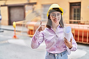 Hispanic girl wearing architect hardhat at construction site smiling happy and positive, thumb up doing excellent and approval