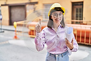 Hispanic girl wearing architect hardhat at construction site smiling with an idea or question pointing finger with happy face,