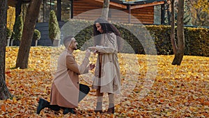 Hispanic loving couple in autumn park man stand in yellow leaves on one knee making marriage proposal girlfriend and