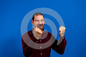 Hispanic man with beard celebrating energetically with clenched fists, isolated on blue studio background.