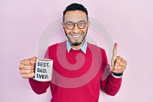 Hispanic man with beard drinking mug of coffee with best dad ever message smiling with an idea or question pointing finger with