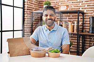 Hispanic man with beard eating delivery salad looking positive and happy standing and smiling with a confident smile showing teeth