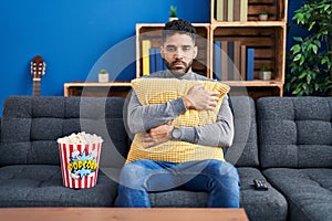 Hispanic man with beard eating popcorn watching a movie at home relaxed with serious expression on face