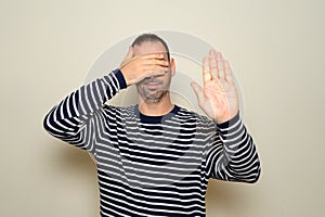 Hispanic man with beard in his 40s wearing a striped sweater covering his eyes with his hands and making stop gesture