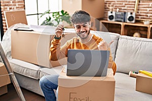 Hispanic man with beard holding keys of new home doing video call with laptop pointing thumb up to the side smiling happy with