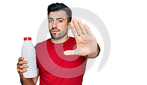 Hispanic man with beard holding liter bottle of milk with open hand doing stop sign with serious and confident expression, defense