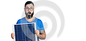 Hispanic man with beard holding photovoltaic solar panel afraid and shocked with surprise and amazed expression, fear and excited