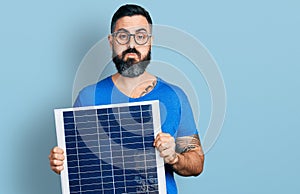 Hispanic man with beard holding photovoltaic solar panel puffing cheeks with funny face