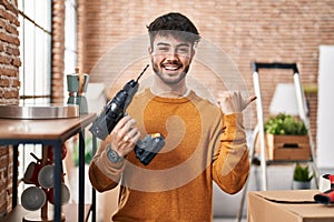 Hispanic man with beard holding screwdriver at new home pointing thumb up to the side smiling happy with open mouth