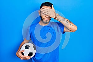 Hispanic man with beard holding soccer ball covering eyes with hand, looking serious and sad