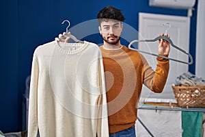Hispanic man with beard holding sweater on hanger at laundry room relaxed with serious expression on face