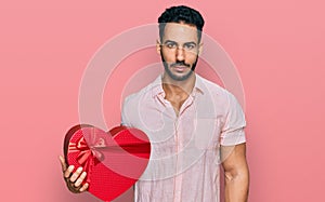 Hispanic man with beard holding valentine gift thinking attitude and sober expression looking self confident